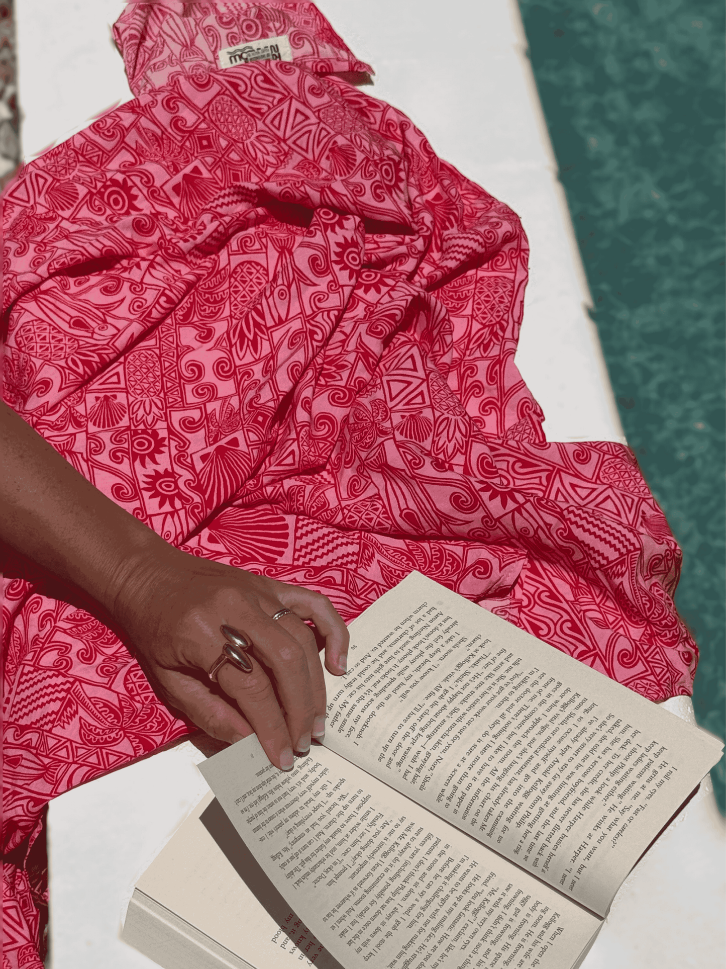 Person wearing a red and pink patterned sarong reading an open book by a poolside.