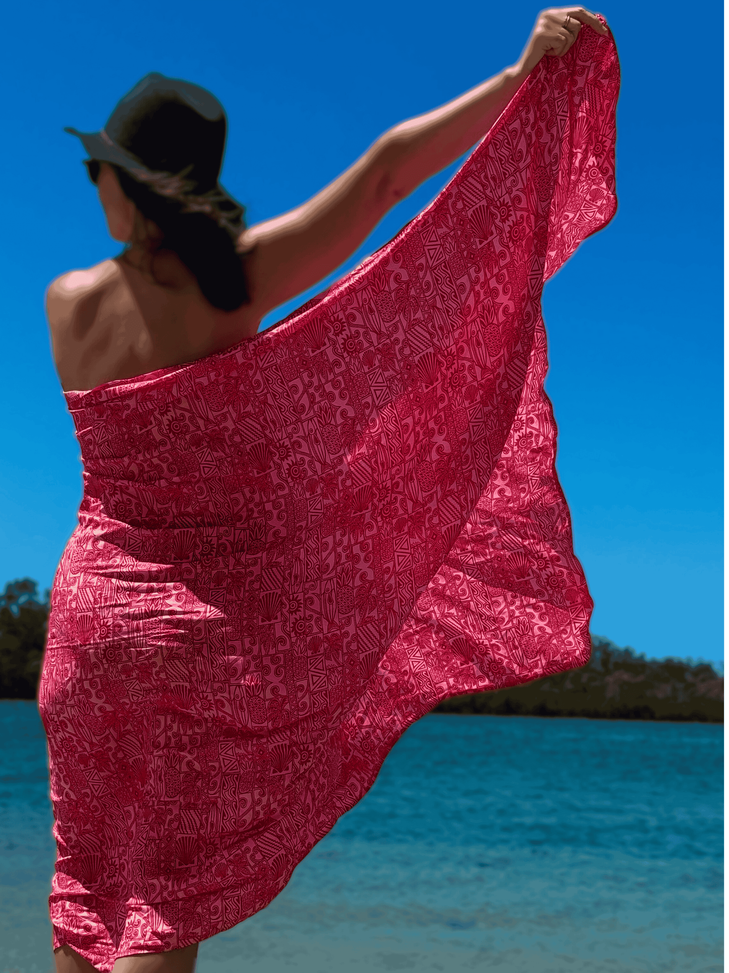 Woman in a red and pink patterned sarong and black hat standing on a beach with clear blue sky.