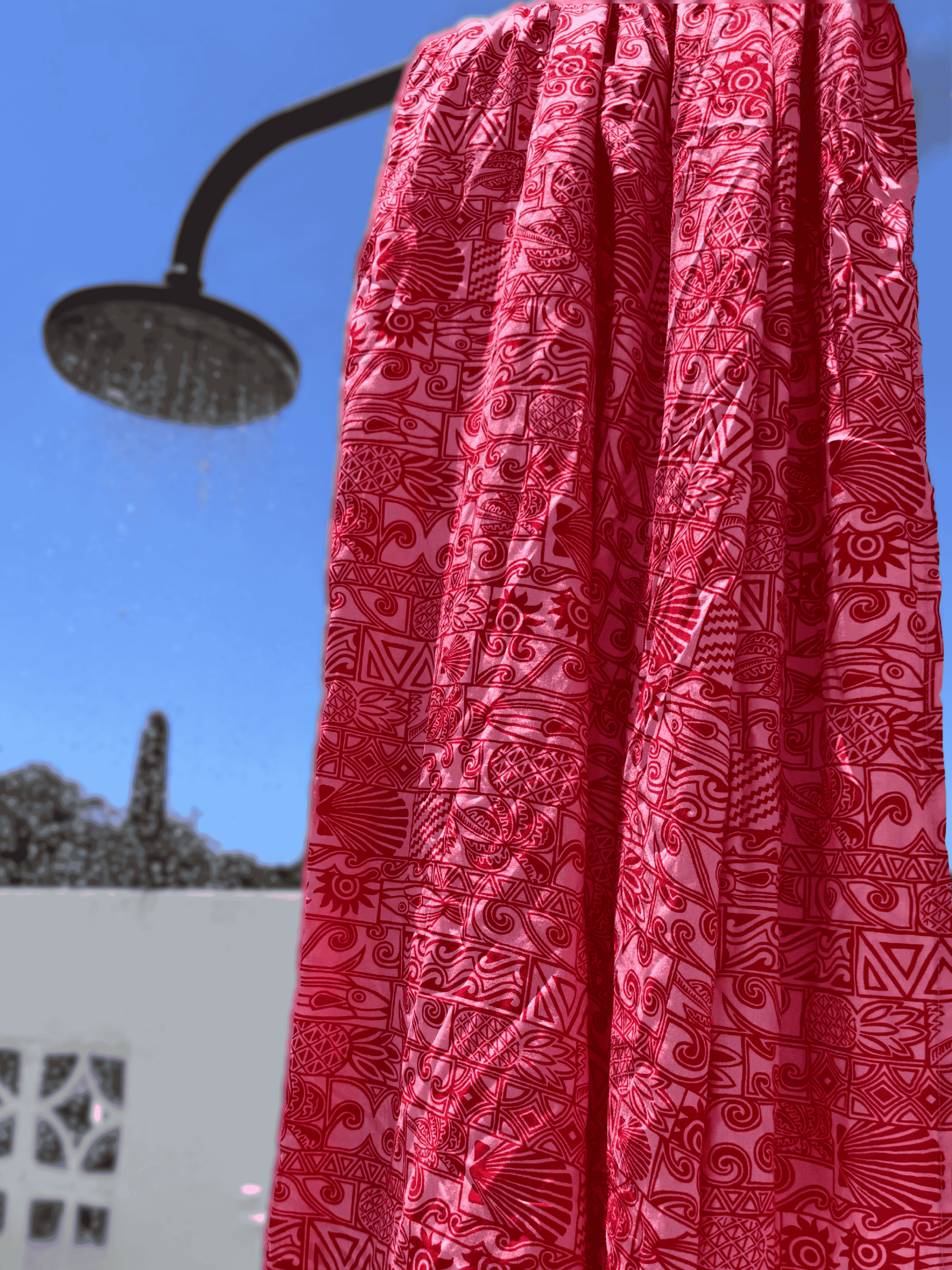 Red and pink patterned sarong hanging from a shower head against a blue sky.
